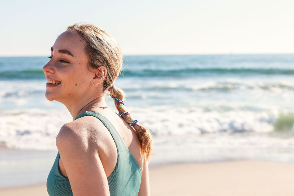 Woman with rip tie mini hair tie in hair standing on a beach with ocean waves in the background
